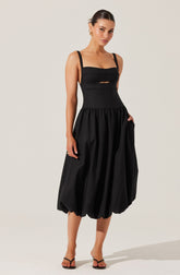A woman is wearing a black sleeveless dress with wide shoulder straps, a fitted bodice with horizontal seam detailing and a small cutout below the chest, and a voluminous, gathered skirt that falls below the knees. She is also wearing black high-heeled sandals and gold earrings.